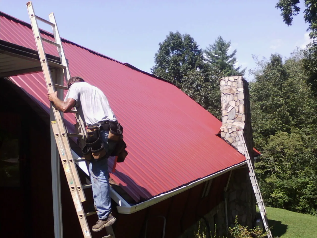 Roofer on a ladder beside a red metal roof and stone chimney — Soffit & Fascia Repair by Roofing Brothers in Linville